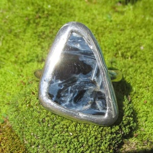 May include: A silver ring with a triangular, polished stone. The stone has a dark blue and black swirling pattern. The ring is set against a background of green moss.