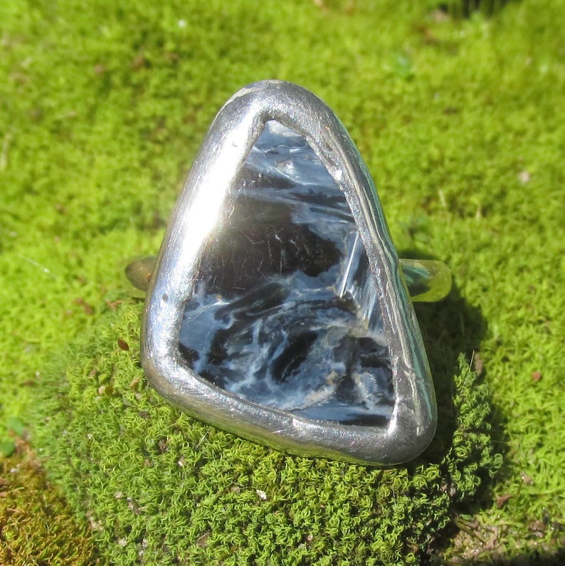 May include: A silver ring with a triangular, polished stone. The stone has a dark blue and black swirling pattern. The ring is set against a background of green moss.