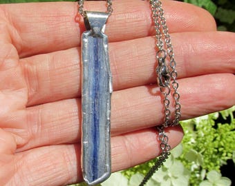 May include: A silver-toned pendant necklace featuring a rectangular, raw-cut blue kyanite stone. The stone has a central band of deep blue, with lighter blue and silver tones. The pendant hangs from a delicate silver chain, creating a natural, elegant accessory.
