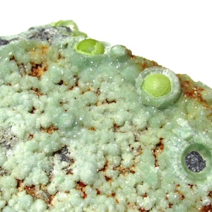 Puede incluir: Primer plano de una muestra mineral verde con una superficie rugosa y texturizada. El mineral tiene una textura porosa y burbujeante y está cubierto de pequeños cristales verdes. Hay varios cristales verdes redondos más grandes en la superficie de la muestra.