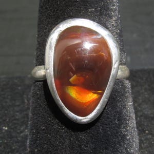 May include: A ring featuring a polished, teardrop-shaped fire agate gemstone set in a smooth, silver-toned metal bezel. The stone displays deep reddish-brown tones with a vibrant, iridescent flash of orange and yellow light across its surface.