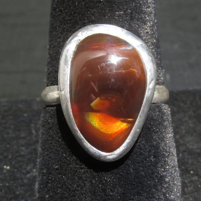 May include: A ring featuring a polished, teardrop-shaped fire agate gemstone set in a smooth, silver-toned metal bezel. The stone displays deep reddish-brown tones with a vibrant, iridescent flash of orange and yellow light across its surface.