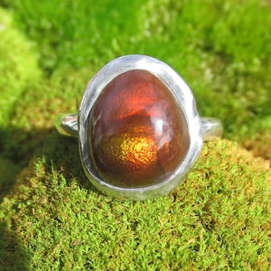 Puede incluir: Un anillo de plata con una piedra preciosa ovalada grande. La piedra tiene un aspecto ardiente e iridiscente, con colores rojizos y dorados. El anillo est&aacute; engastado en una banda de plata pulida y sencilla. Se muestra sobre musgo verde.