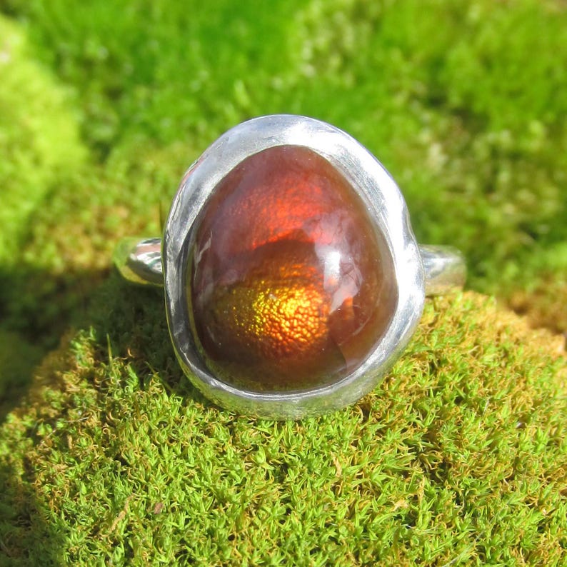 Puede incluir: Un anillo de plata con una piedra preciosa ovalada grande. La piedra tiene un aspecto ardiente e iridiscente, con colores rojizos y dorados. El anillo est&aacute; engastado en una banda de plata pulida y sencilla. Se muestra sobre musgo verde.