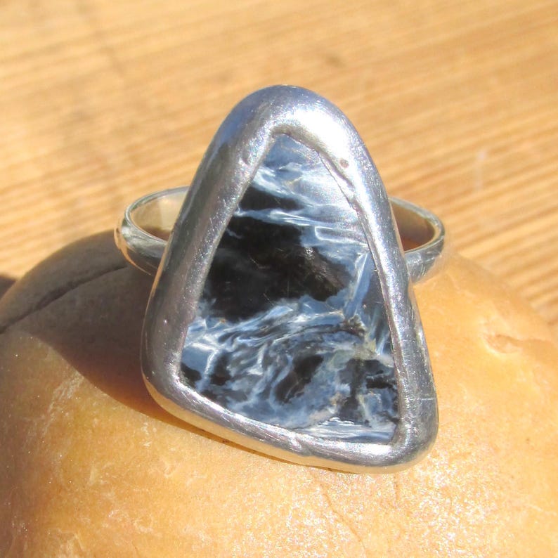 May include: A silver ring featuring a triangular, polished gemstone with a swirling pattern of blue and black. The stone is set in a silver bezel, creating a striking contrast. The ring is displayed on a light brown surface.
