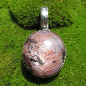 May include: A round pendant featuring a polished, mottled stone with shades of dusty pink, reddish-brown, and dark charcoal inclusions. The stone is set in a smooth, silver-toned metal bezel with a matching silver-toned metal bail at the top.