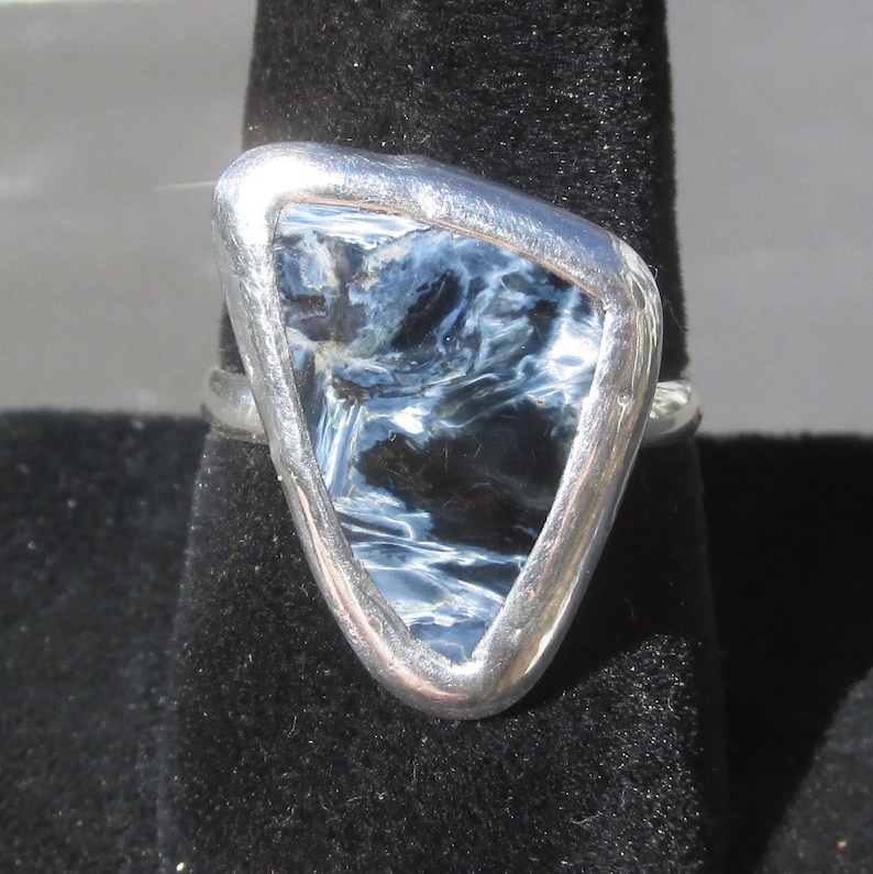 May include: A silver ring featuring a triangular, polished gemstone with swirling patterns of blue and black. The stone is set in a silver bezel, creating a modern and elegant design. The ring is displayed on a black velvet surface.