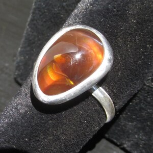 May include: A silver ring featuring a large, smooth, teardrop-shaped fire opal gemstone. The stone displays deep orange and reddish-brown hues with internal flashes of yellow and green light, set within a simple, polished silver bezel and band.