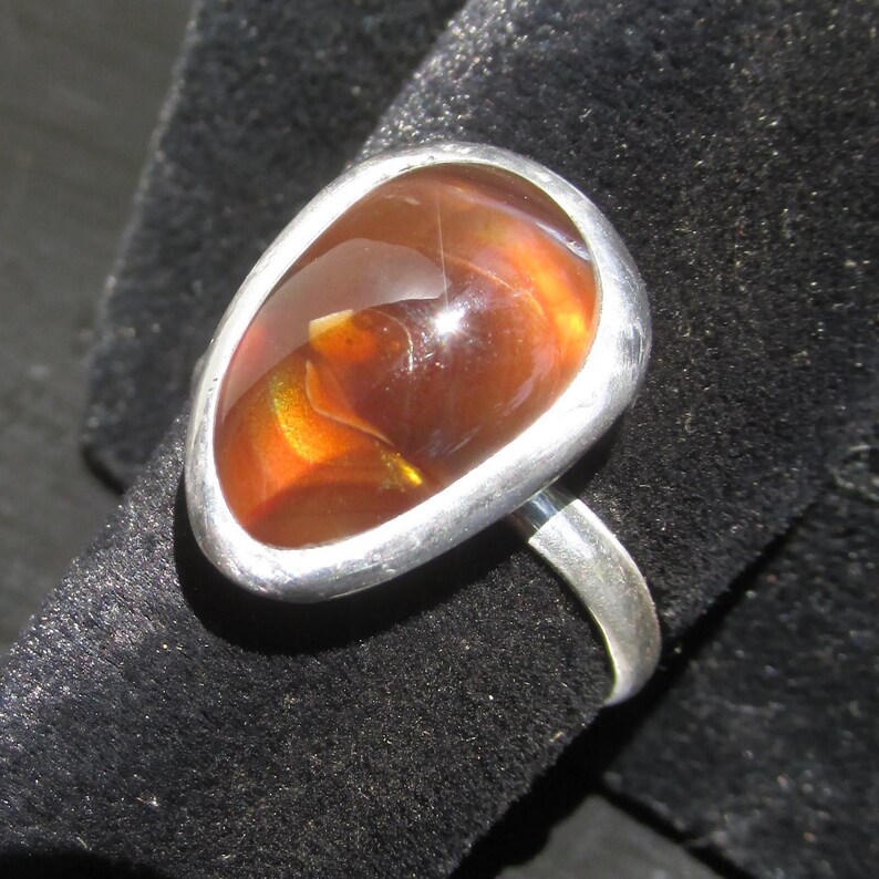 May include: A silver ring featuring a large, smooth, teardrop-shaped fire opal gemstone. The stone displays deep orange and reddish-brown hues with internal flashes of yellow and green light, set within a simple, polished silver bezel and band.