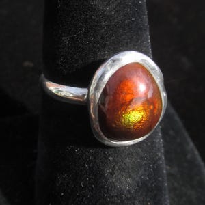 Puede incluir: Un anillo de plata con una piedra preciosa grande, de forma ovalada, con un aspecto iridiscente y ardiente. Los colores de la piedra cambian de rojos y naranjas profundos a verdes y amarillos vibrantes. El anillo est&aacute; engastado en una banda de plata pulida.