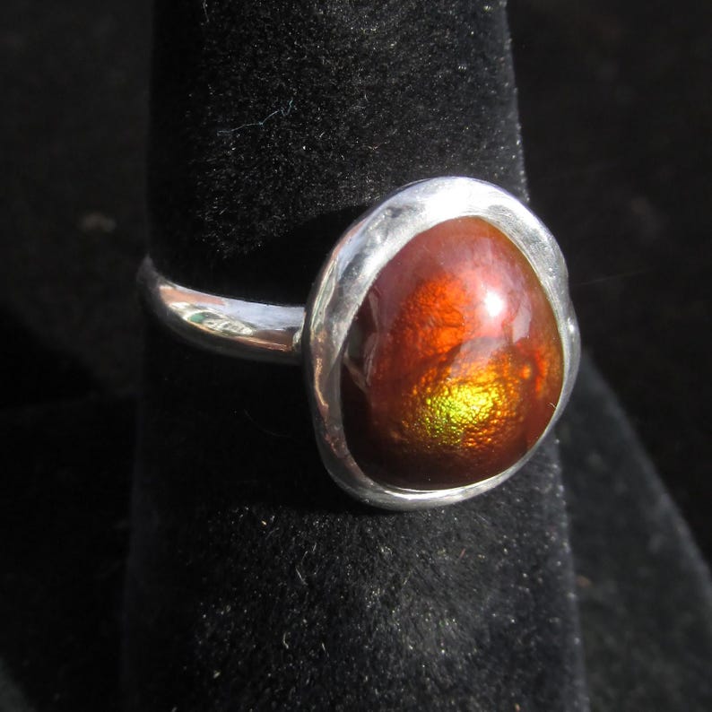 Puede incluir: Un anillo de plata con una piedra preciosa grande, de forma ovalada, con un aspecto iridiscente y ardiente. Los colores de la piedra cambian de rojos y naranjas profundos a verdes y amarillos vibrantes. El anillo est&aacute; engastado en una banda de plata pulida.