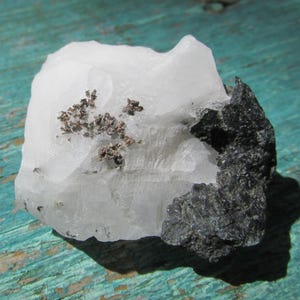 May include: A raw mineral specimen featuring a large, translucent white quartz crystal formation. Embedded within the white quartz are small, metallic silver-colored dendritic inclusions, accompanied by a dark, rough, charcoal-gray rock mass.
