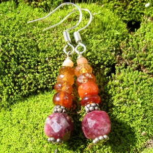 May include: Dangling earrings with a silver hook and a beaded design. The beads are a gradient of colors, starting with light peach, transitioning to amber and orange, and ending with a large, faceted, deep red bead. The earrings are set against a green moss background.