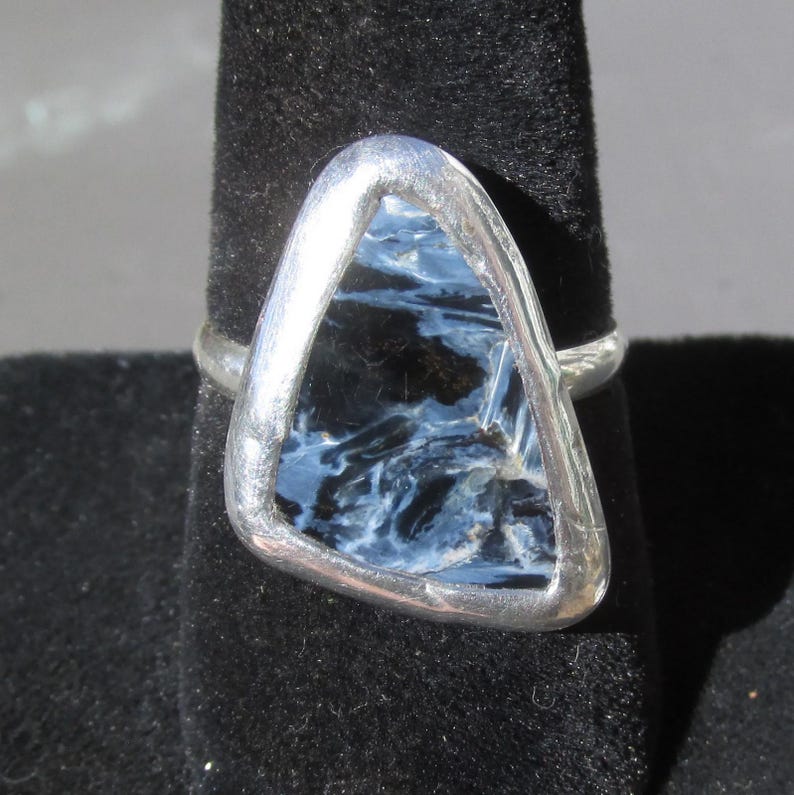 May include: A silver ring featuring a triangular, polished gemstone with a swirling pattern of blue and black hues. The stone is set in a silver bezel, creating a modern and elegant design. The ring is displayed on a black surface.