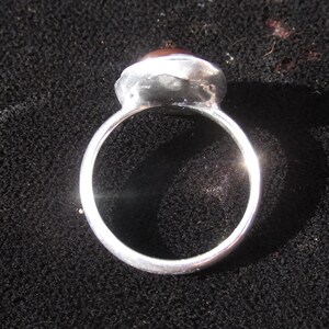 Puede incluir: Un anillo de plata con una banda redonda y una piedra grande, ovalada, de color marr&oacute;n rojizo. La piedra est&aacute; engastada en un bisel de plata. El anillo se muestra sobre una superficie negra.