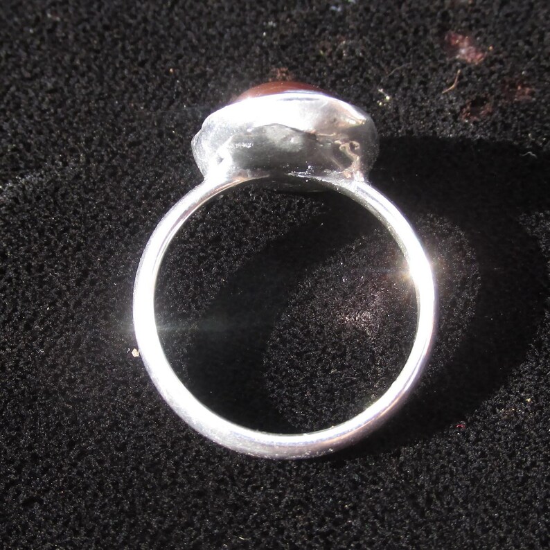 Puede incluir: Un anillo de plata con una banda redonda y una piedra grande, ovalada, de color marr&oacute;n rojizo. La piedra est&aacute; engastada en un bisel de plata. El anillo se muestra sobre una superficie negra.