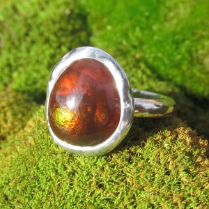 Puede incluir: Un anillo de plata con una piedra preciosa ovalada grande. La piedra es una mezcla de marr&oacute;n y naranja con reflejos verdes. El anillo est&aacute; sobre musgo verde. El dise&ntilde;o del anillo es simple y elegante.