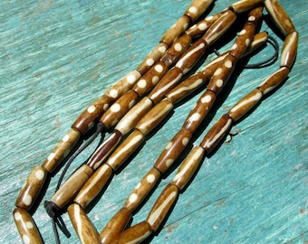 May include: A necklace featuring elongated, brown beads with white dotted and striped patterns. The beads are strung on a black cord, creating a rustic and earthy aesthetic. The necklace is displayed on a turquoise surface.