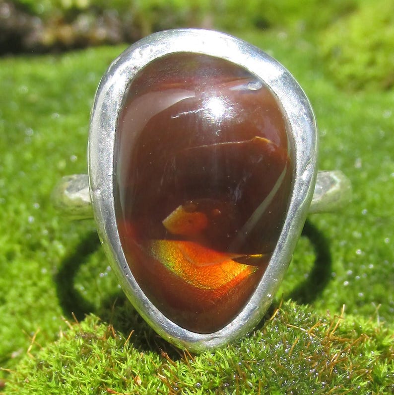 May include: A silver ring featuring a large, teardrop-shaped, dark brown fire opal gemstone. The stone displays a vibrant, iridescent orange and yellow flash within its polished, translucent surface. The metal band has a hammered texture.