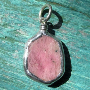 May include: A pendant featuring a pink gemstone encased in a silver-colored metal frame. The gemstone has a textured appearance with varying shades of pink. The pendant includes a silver loop for hanging.