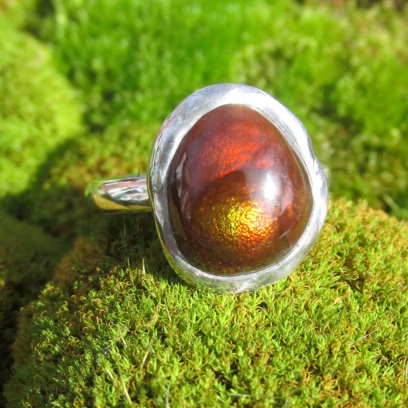 Puede incluir: Un anillo de plata con una gran gema ovalada en tonos marr&oacute;n, naranja y dorado. El anillo est&aacute; sobre un lecho de musgo verde. Los colores de la gema son vibrantes, creando un contraste llamativo.
