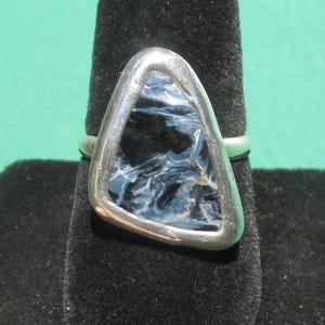 May include: A silver ring featuring a triangular, polished gemstone with a swirling pattern of dark blue and white. The stone is set within a silver bezel, creating a modern and elegant design. The ring is displayed on a black surface.