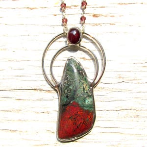 Puede incluir: Collar con colgante de plata con una piedra grande de forma irregular en tonos verdes y rojos. Encima, una piedra roja redonda más pequeña en un halo plateado. La cadena del collar tiene pequeñas cuentas rojas.