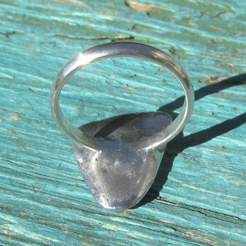 May include: A silver ring with a teardrop-shaped setting. The ring is set against a turquoise wooden surface, with the sunlight casting a shadow. The ring's band is smooth and rounded.