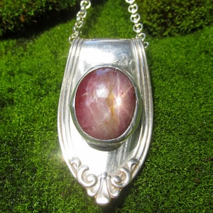 May include: A silver pendant necklace featuring a polished, oval-shaped cabochon star ruby set in the center of a decorative, vintage-style silver spoon handle. The pendant hangs from a delicate silver chain, showcasing a smooth, metallic finish.