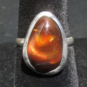 May include: A silver ring featuring a large, teardrop-shaped fire agate gemstone. The stone displays deep reddish-brown tones with a bright, iridescent orange and yellow flash in the center. The metal setting is smooth, polished, and organic in shape.