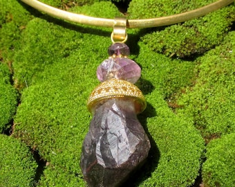 May include: A gold-toned metal collar necklace featuring a pendant with a faceted purple amethyst crystal point. The pendant is accented by a smaller, rounded purple amethyst bead and a decorative gold-toned metal cap with a textured, beaded edge.