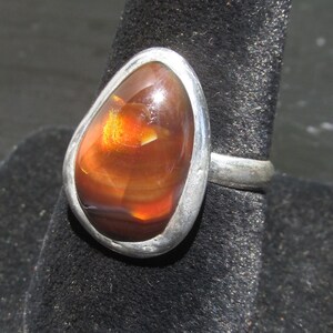 May include: A silver ring featuring a large, teardrop-shaped fire agate gemstone. The stone displays deep reddish-brown tones with a bright, glowing orange-gold flash within its polished, smooth surface, set in a simple, solid silver bezel.