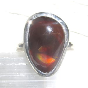 May include: A ring featuring a large, teardrop-shaped dark brown stone set in a smooth, polished silver-toned metal band. The stone displays internal flashes of iridescent orange and green light, with a glossy, reflective surface finish.