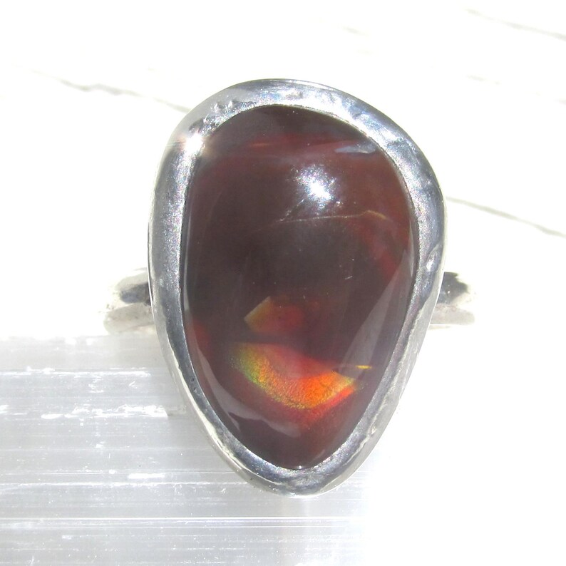 May include: A ring featuring a large, teardrop-shaped dark brown stone set in a smooth, polished silver-toned metal band. The stone displays internal flashes of iridescent orange and green light, with a glossy, reflective surface finish.