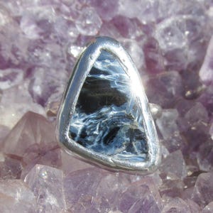 May include: A silver ring featuring a triangular, polished gemstone with a swirling pattern of blue and black. The ring is set against a backdrop of purple amethyst crystals, creating a striking contrast.