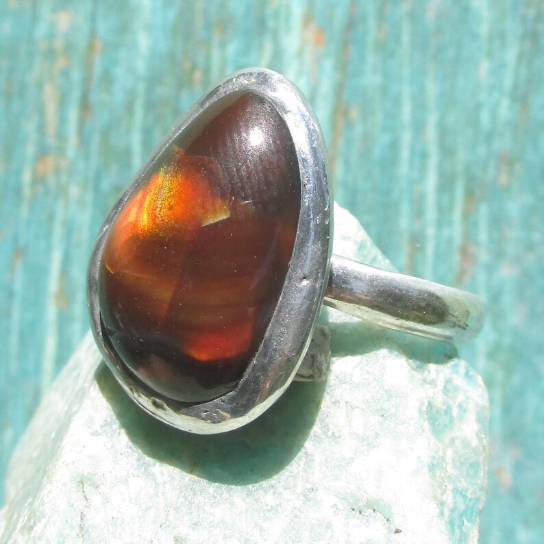 May include: A silver ring featuring a teardrop-shaped fire agate gemstone. The stone displays deep brown tones with a vibrant, glowing orange and red iridescent flash. The metal band is smooth and polished, securely holding the cabochon stone.