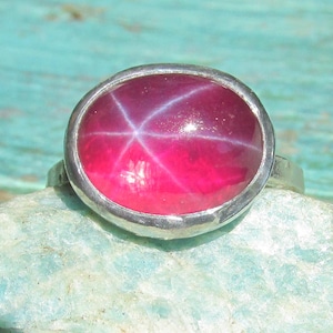 May include: A silver ring featuring a large, oval-shaped, deep pink star ruby cabochon. The gemstone displays a distinct six-rayed star pattern across its smooth, polished surface, set within a simple, polished silver bezel and band.