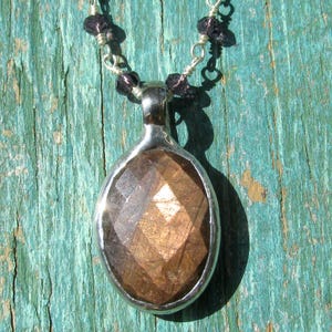 Könnte beinhalten: Eine silberne Anhängerkette mit einem facettierten, braunen Edelstein. Der ovale Anhänger hat eine silberne Lünette. Die Halskette besteht aus kleinen, dunkelvioletten Perlen und Silberdraht.