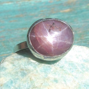 Pode incluir: Um anel de prata com um grande rubi estrela de corte cabochão oval. A pedra preciosa exibe um efeito de estrela de seis raios na sua superfície translúcida de cor rosa-púrpura profundo, engastada numa moldura de prata lisa e polida.
