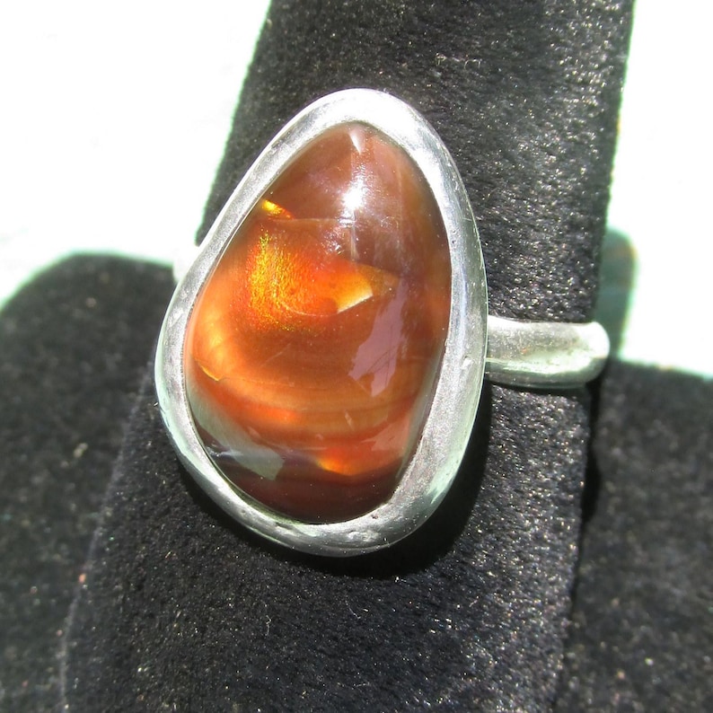 May include: A silver ring featuring a smooth, teardrop-shaped fire agate gemstone. The stone displays deep reddish-brown tones with a bright, glowing orange iridescent flash in the center, set within a polished, solid silver bezel setting.