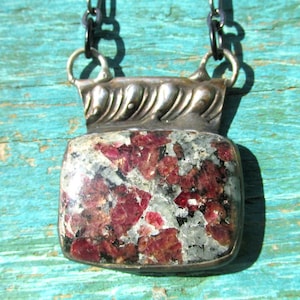 May include: A silver pendant necklace with a rectangular stone displaying red and grey tones. The stone is set in a decorative silver frame and suspended from a dark chain. The pendant measures approximately 3.8 cm by 2.5 cm.