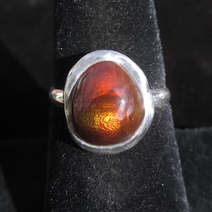 Puede incluir: Un anillo de plata con una piedra preciosa grande, de forma ovalada, con un aspecto &iacute;gneo e iridiscente. La piedra muestra una mezcla de tonos naranja, rojo y dorado, engastada en una banda de plata pulida y sencilla. El anillo se muestra sobre un fondo oscuro.