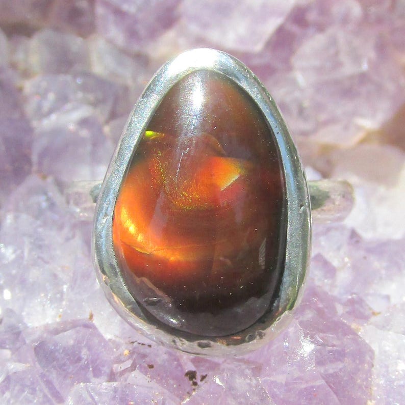May include: A teardrop-shaped fire opal ring set in a smooth silver bezel. The gemstone displays a deep reddish-brown base color with vibrant flashes of orange and yellow iridescence across its polished, domed surface, mounted on a simple silver band.