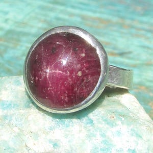 May include: A silver ring featuring a large, round, cabochon-cut deep magenta ruby gemstone set in a smooth metal bezel. The stone displays natural inclusions and a polished surface, while the band is made of a plain, solid silver metal.