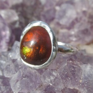 Puede incluir: Anillo de plata con una piedra preciosa ovalada. La piedra tiene un aspecto ardiente e iridiscente, con tonos rojos, naranjas y verdes. El anillo est&aacute; sobre una superficie p&uacute;rpura.