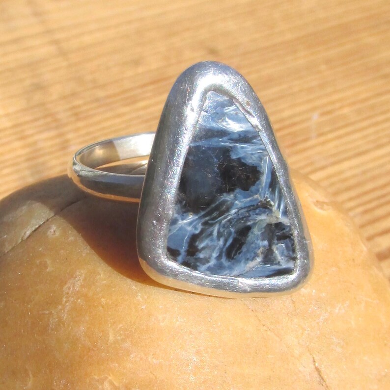 May include: A silver ring featuring a triangular, polished gemstone with swirling patterns of blue and black. The stone is set in a silver bezel, creating a striking contrast. The ring is displayed on a light brown surface.