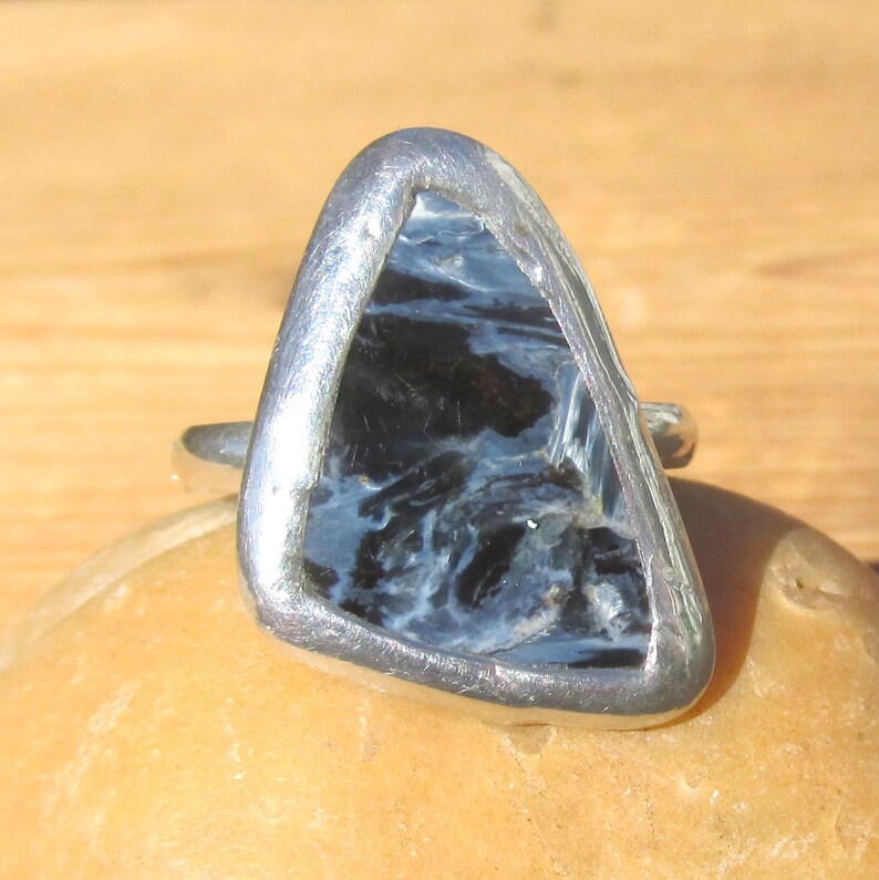 May include: A silver ring featuring a triangular, polished gemstone with a swirling pattern of blue and black. The stone is set in a silver bezel, creating a modern and unique design. The ring is displayed on a light brown surface.