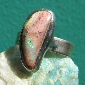 May include: A silver ring featuring an oval-shaped opal gemstone. The opal displays a mix of colors, including pink, white, and hints of green and blue. The ring's band is a simple, polished silver.