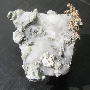 May include: A natural mineral specimen with a white quartz base, adorned with clusters of grey and copper-coloured formations. The rough, irregular shape of the stone highlights its raw beauty. The copper-coloured section provides a striking contrast.