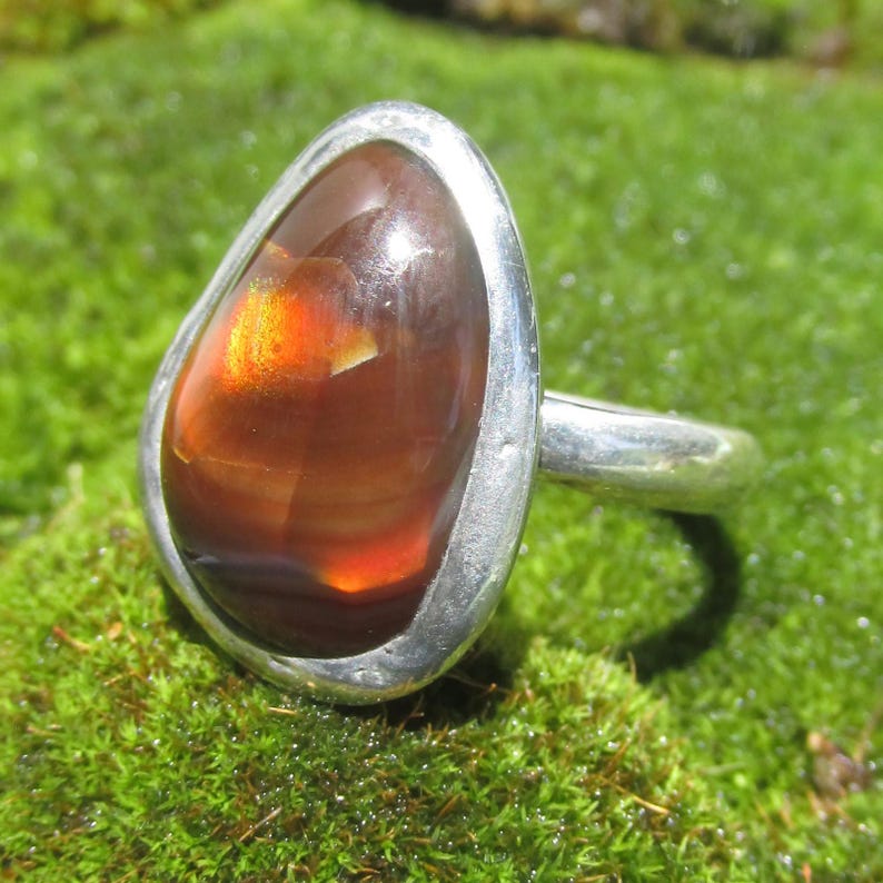 May include: A silver ring featuring a smooth, teardrop-shaped fire opal gemstone. The stone displays a deep reddish-brown base color with a bright, glowing orange internal flash. The metal band is polished and holds the stone in a bezel setting.